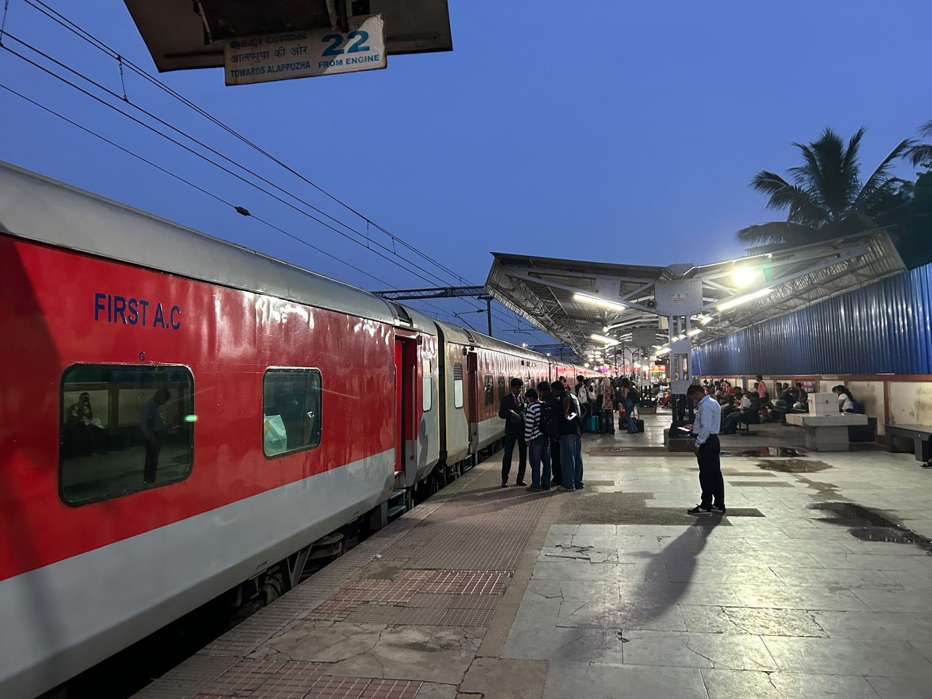 A red and silver First A.C. railway carriage at an illuminated railway platform at dusk
